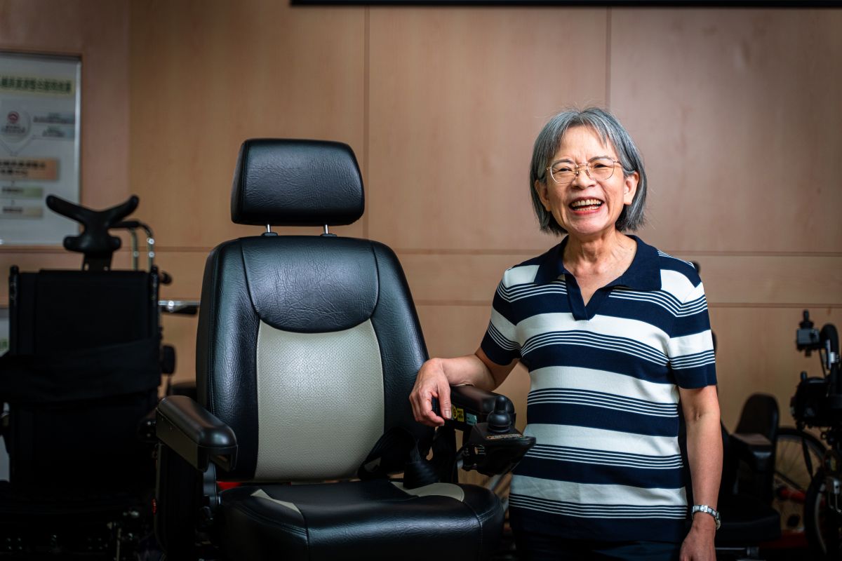 A smiling woman standing next to a powered mobility chair inside an assistive technology display area.