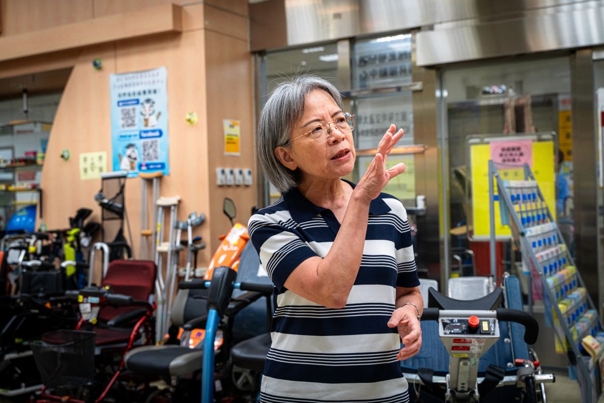 A woman speaking and gesturing inside an assistive device exhibition space, surrounded by various mobility and assistive equipment.